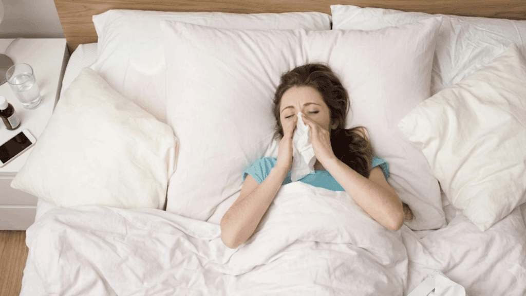 Woman in blue shirt in bed, blowing nose with a tissue.
