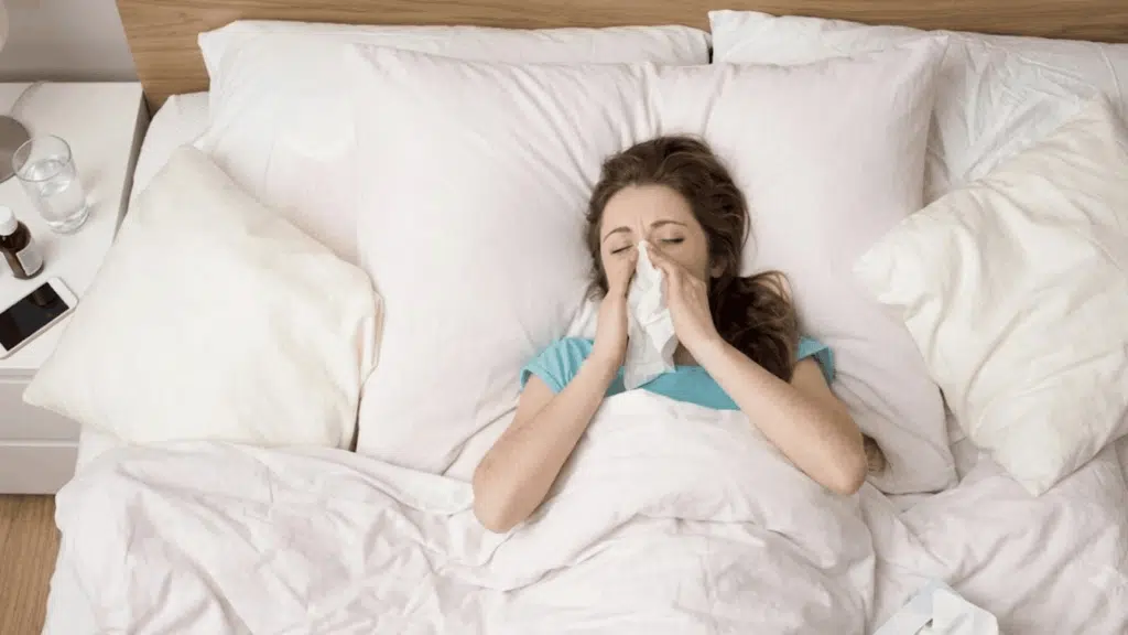 Woman in blue shirt in bed, blowing nose with a tissue.