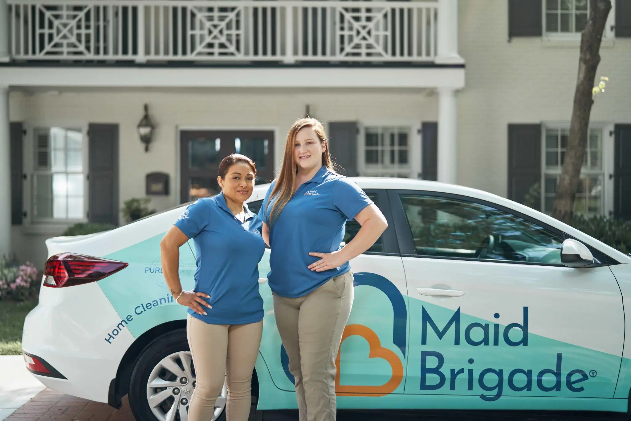 Two women in blue uniforms standing beside Maid Brigade car outside residential home