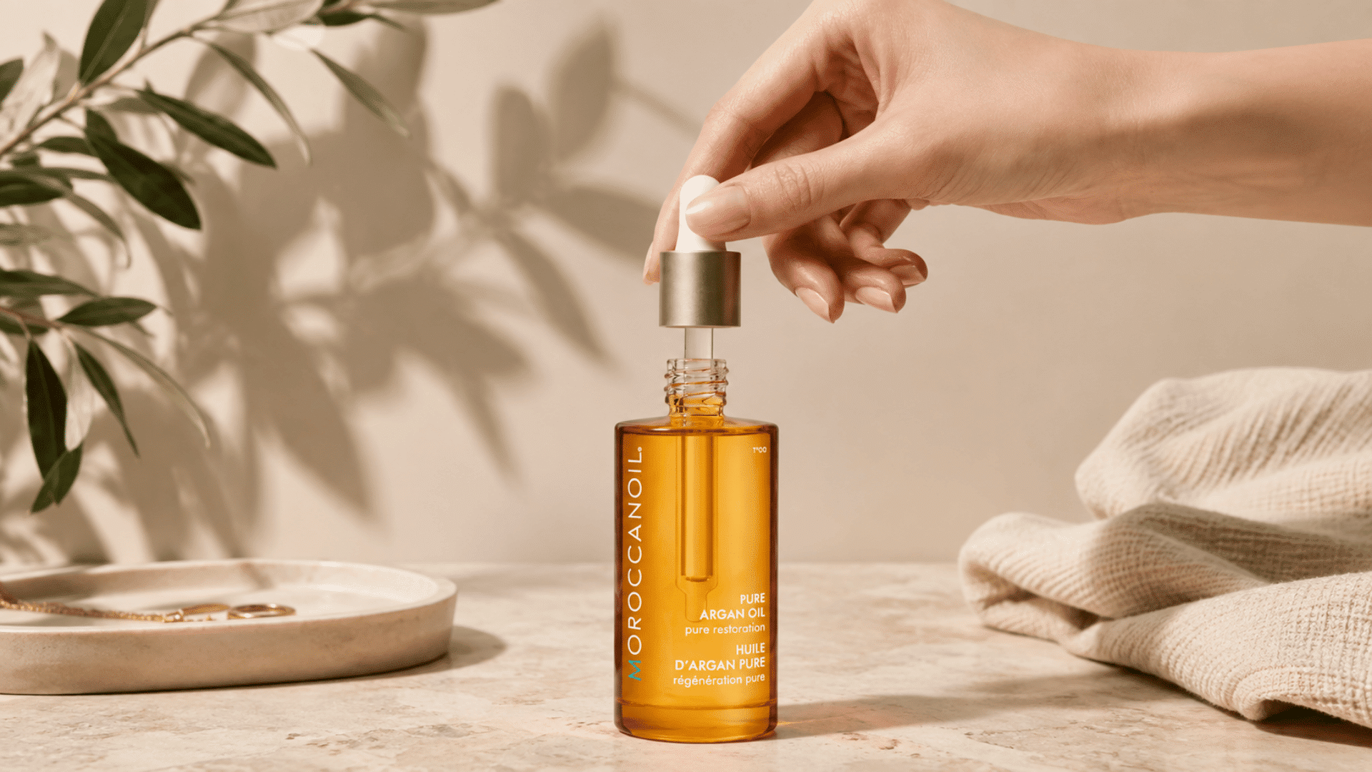 Hand holding a dropper above a Moroccanoil Pure Argan Oil bottle on a neutral stone surface with greenery and soft spa décor.