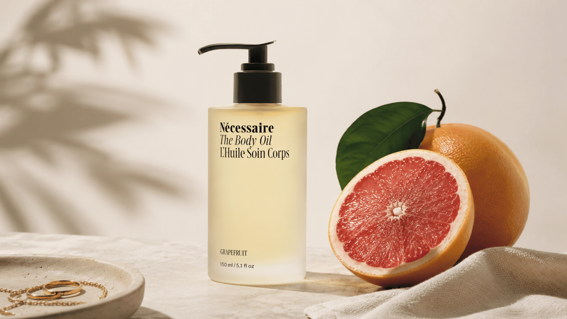 Nécessaire The Body Oil bottle displayed beside fresh grapefruit on a neutral surface with soft natural shadows.