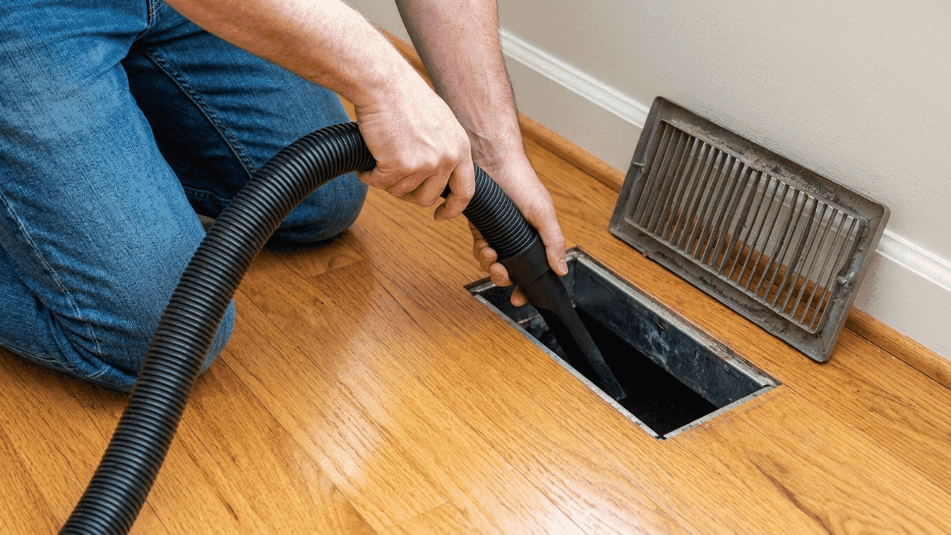 person cleaning an open floor air duct with a vacuum hose beside the vent cover