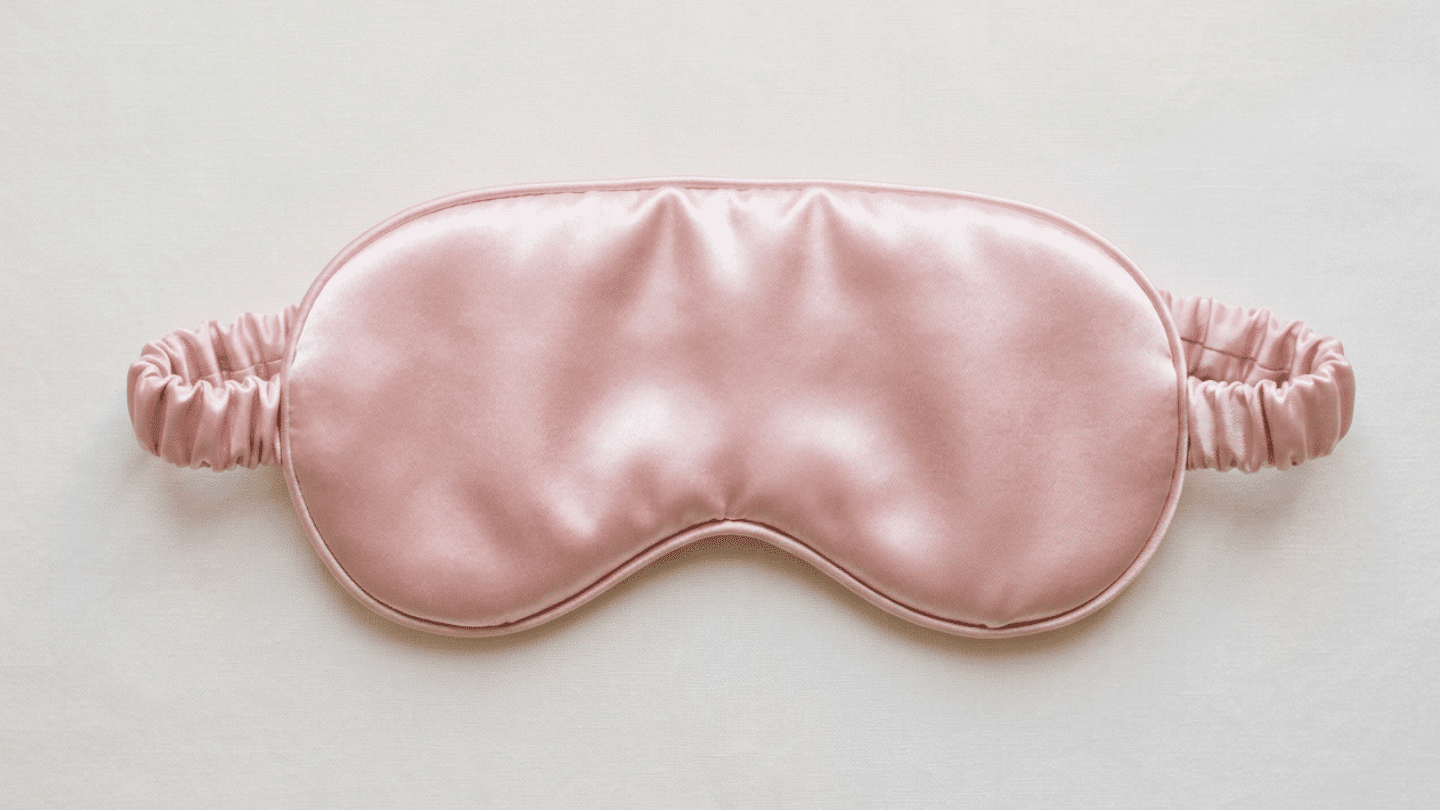 pink Eberjey satin sleep mask with soft padding and a covered elastic band on white fabric laid flat