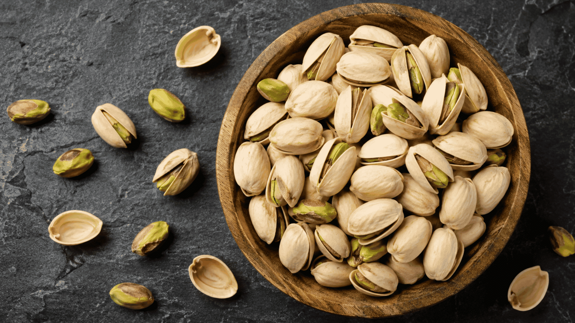 pistachios in shells and kernels arranged in a wooden bowl on dark stone