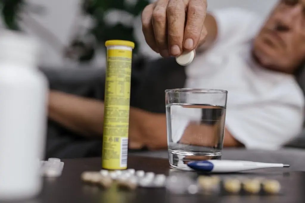 Person dropping tablet into glass of water surrounded by medication on table