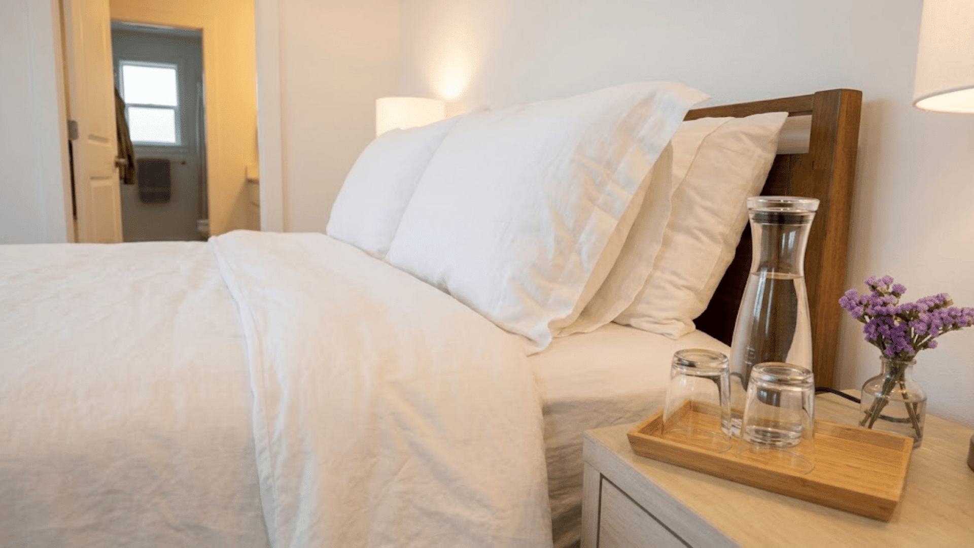 Clear water carafe and two glasses on a wooden tray atop a guest nightstand.