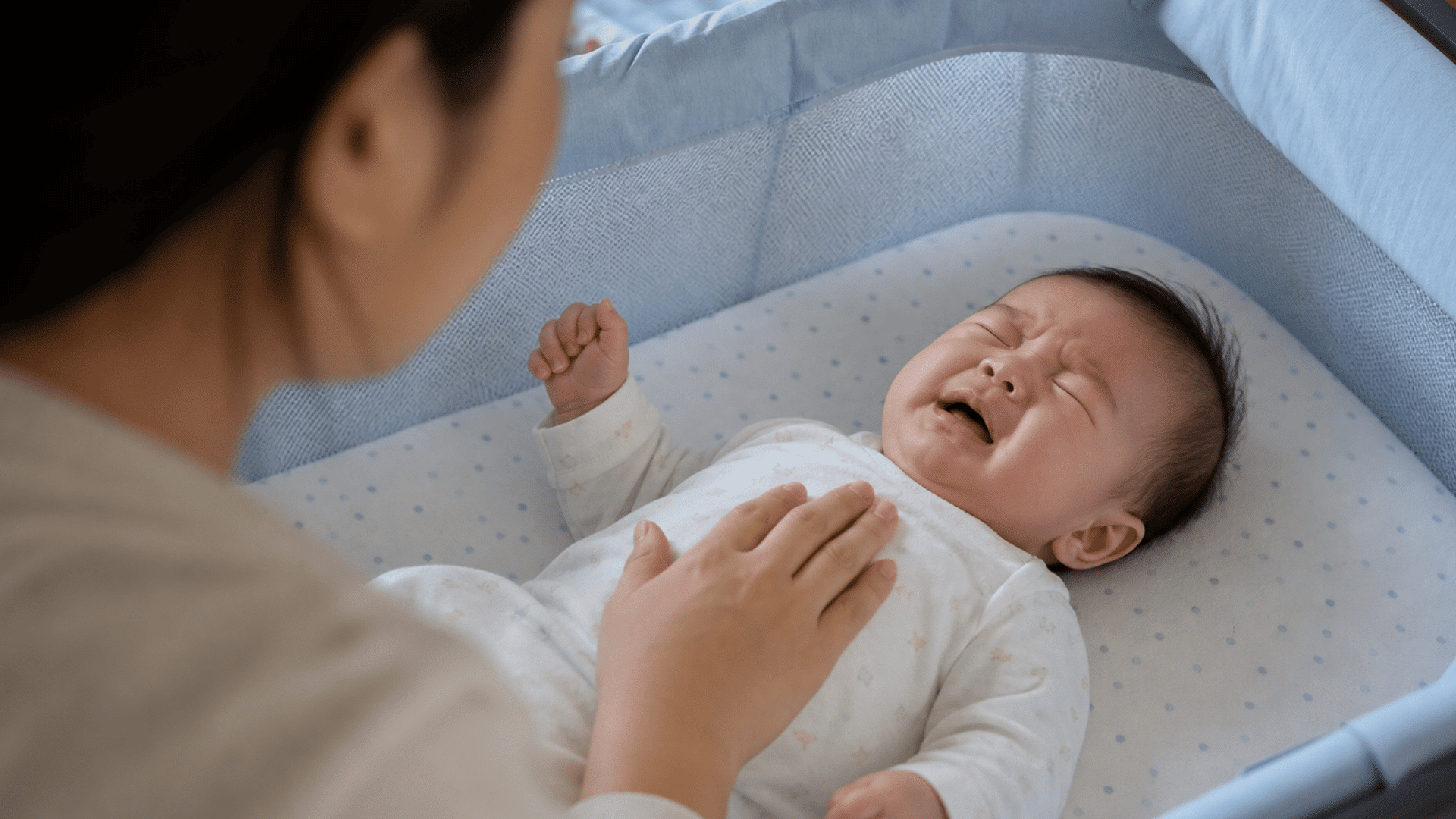 baby crying with eyes closed in a bassinet while a parent gently comforts the baby.