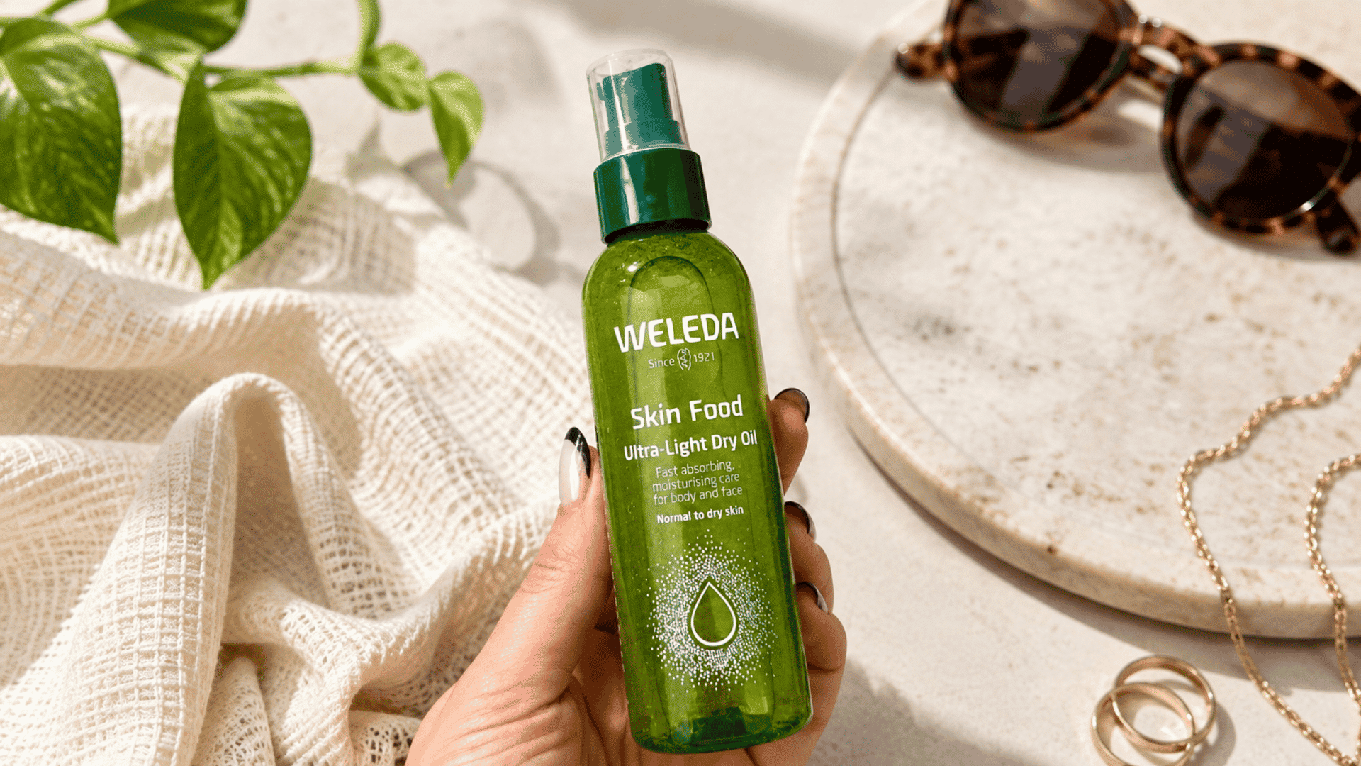 Weleda Skin Food Ultra-Light Dry Oil bottle held over a sunlit surface with sunglasses, greenery, and gold accessories.