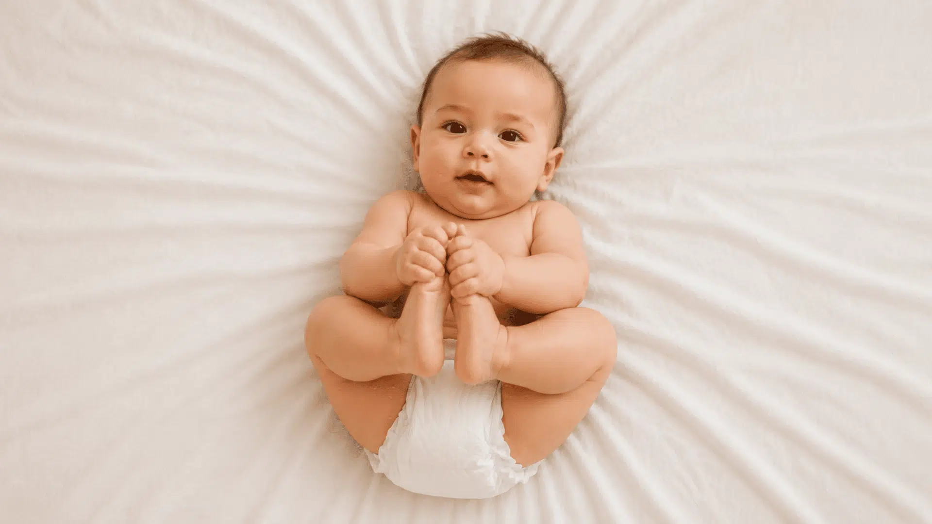 baby in a diaper lying on a white bed sheet holding its own feet while smiling up