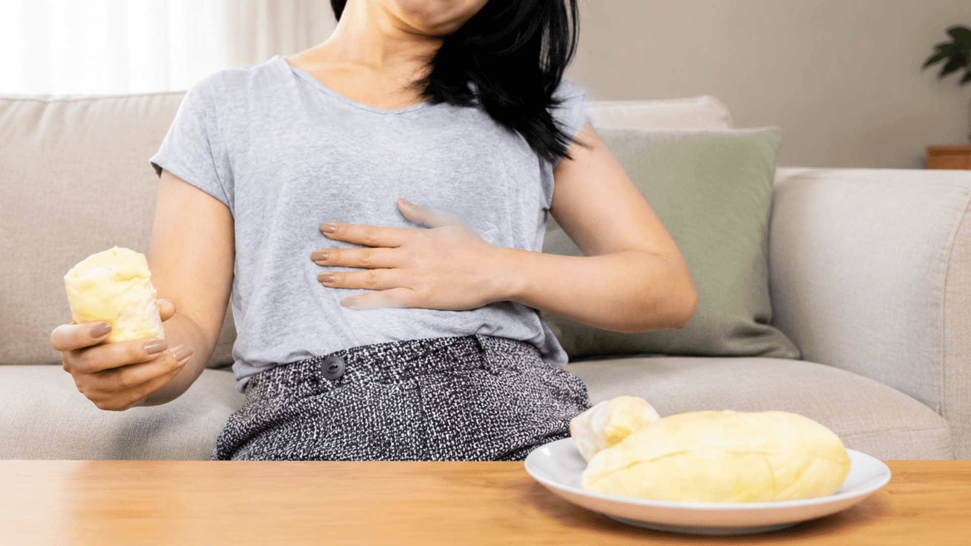 Woman experiencing digestive discomfort while holding fruit beside a plate on a table.