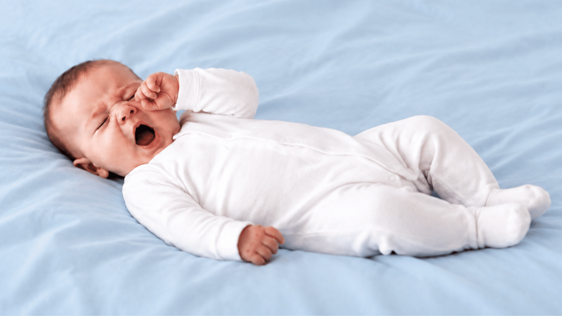 Yawning baby in white pajamas lying on a light blue bed.
