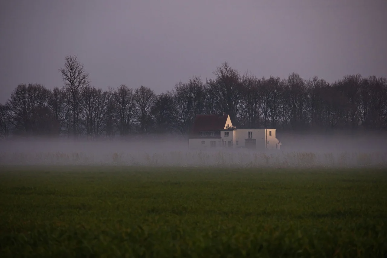 House surrounded by fog in a field with trees in the background at dusk