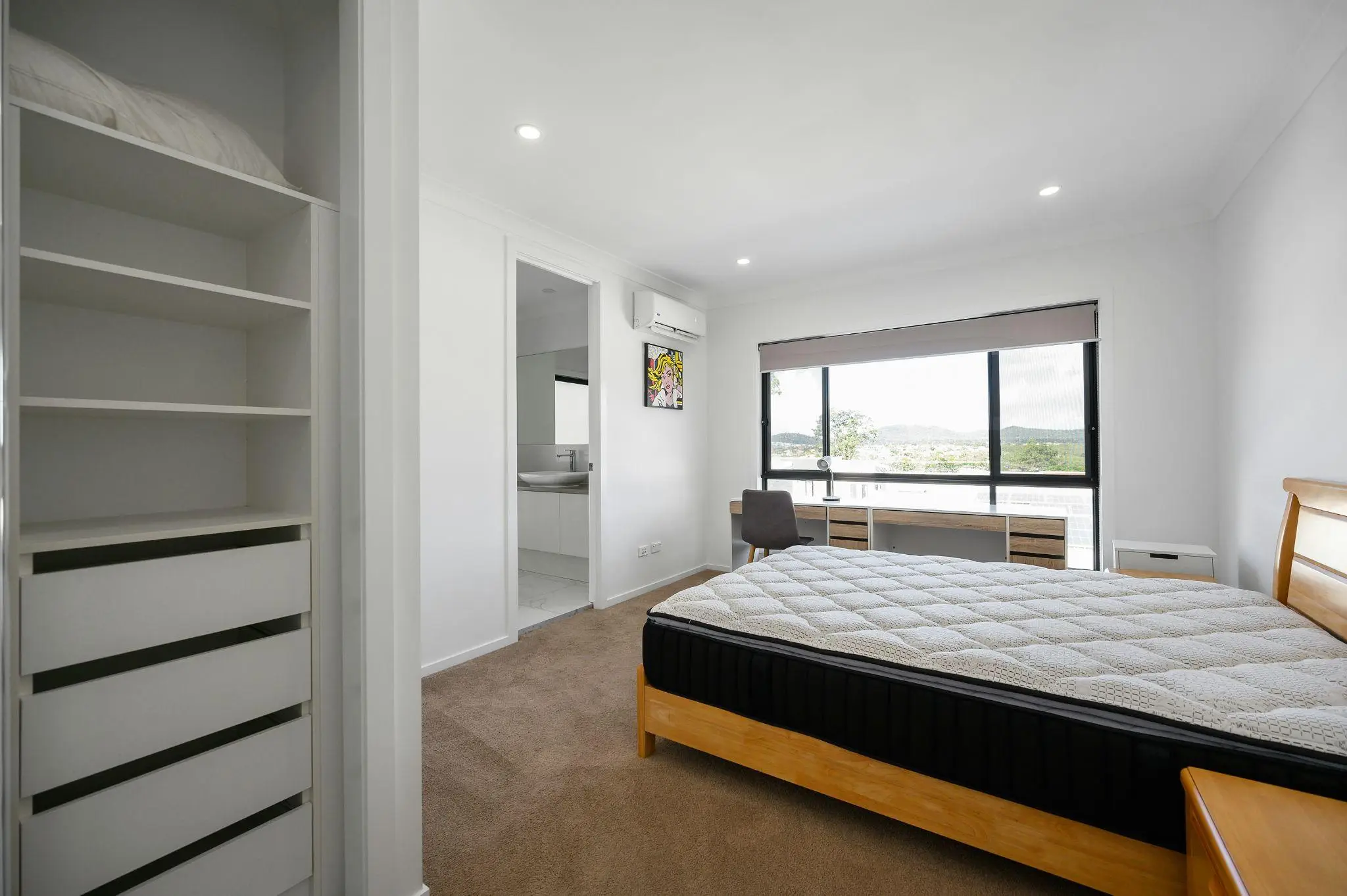 Modern bedroom with wooden bedframe, desk near large window, white walls, and an open closet