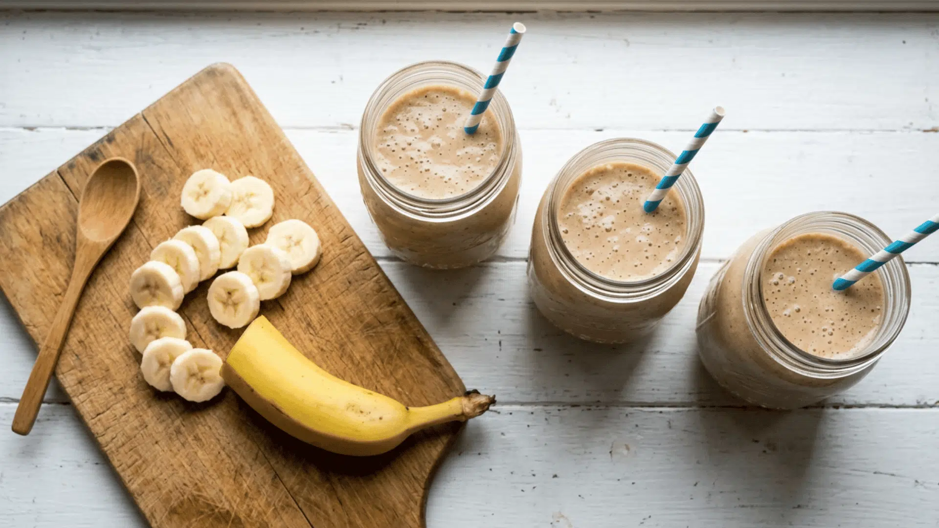 three banana protein shakes with striped straws beside sliced banana on a wooden board