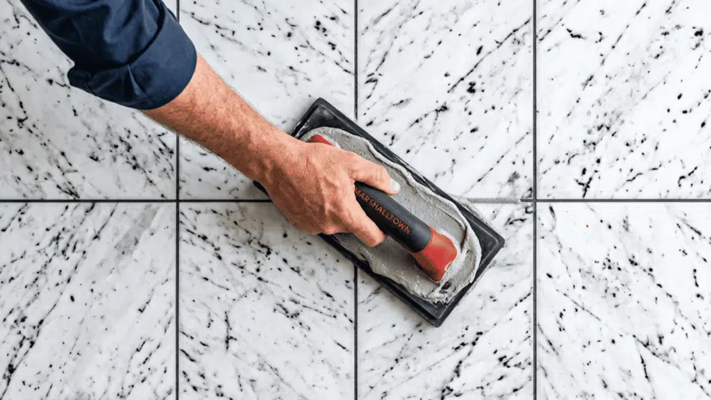 worker using a grout float on white marble-look floor tiles with fresh gray grout lines between the joints