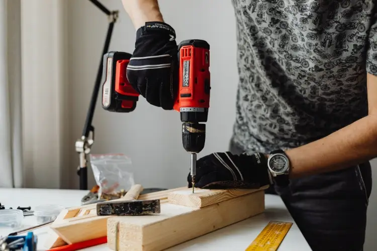 Person using red cordless drill on wooden planks in home workshop with scattered tools