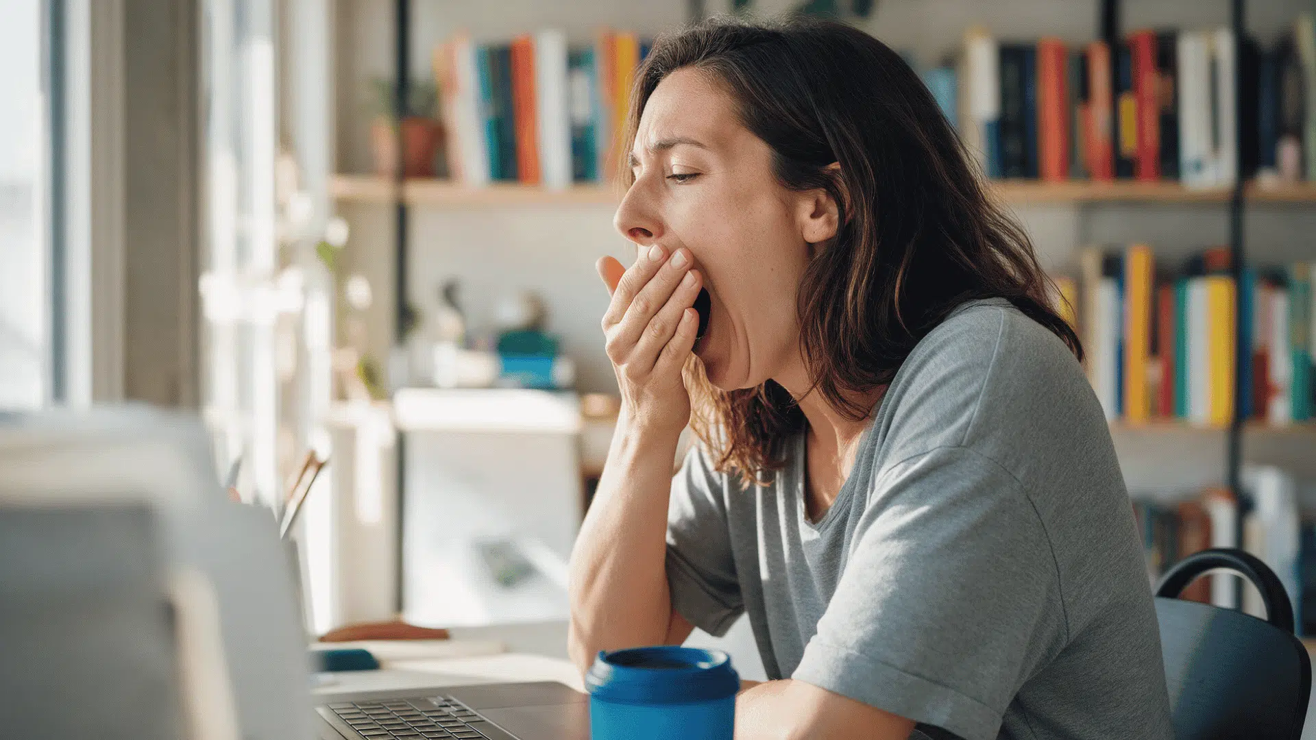 woman yawning while working on a laptop at home with bookshelves in the background
