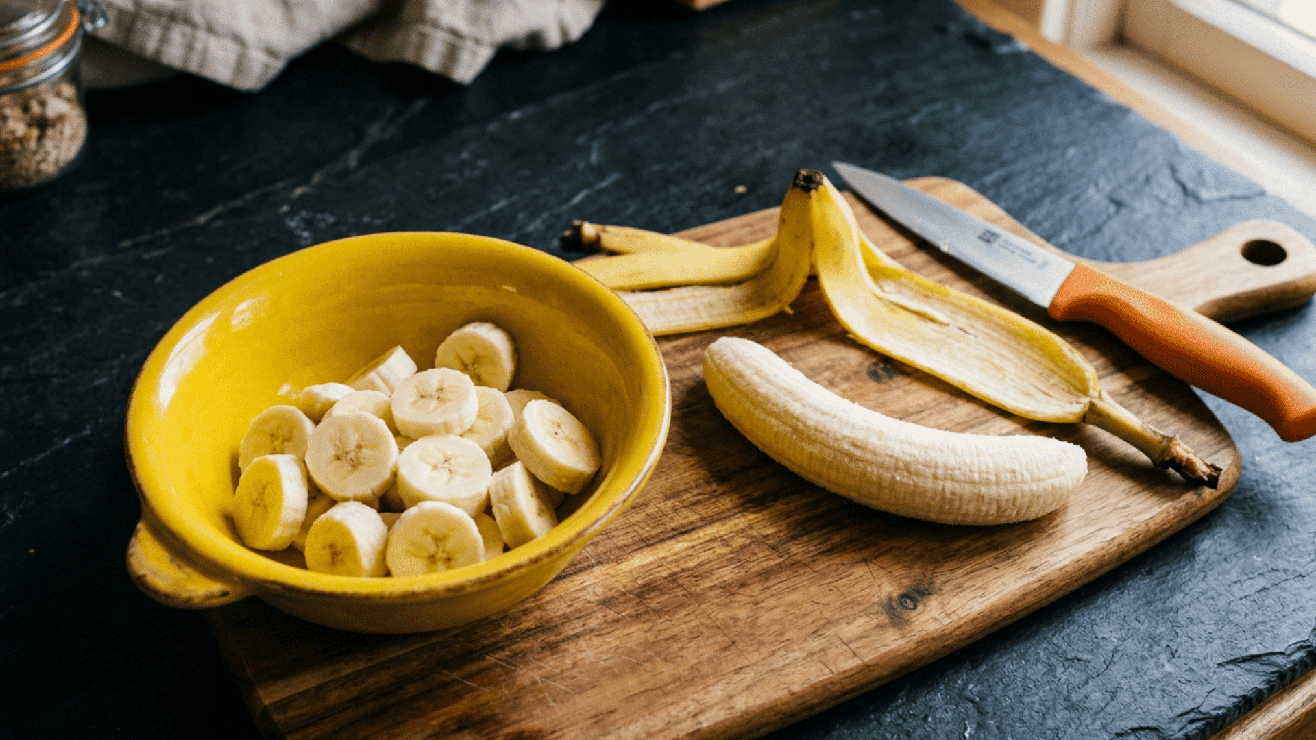 sliced banana in a yellow bowl beside a peeled banana and knife on a wooden cutting board