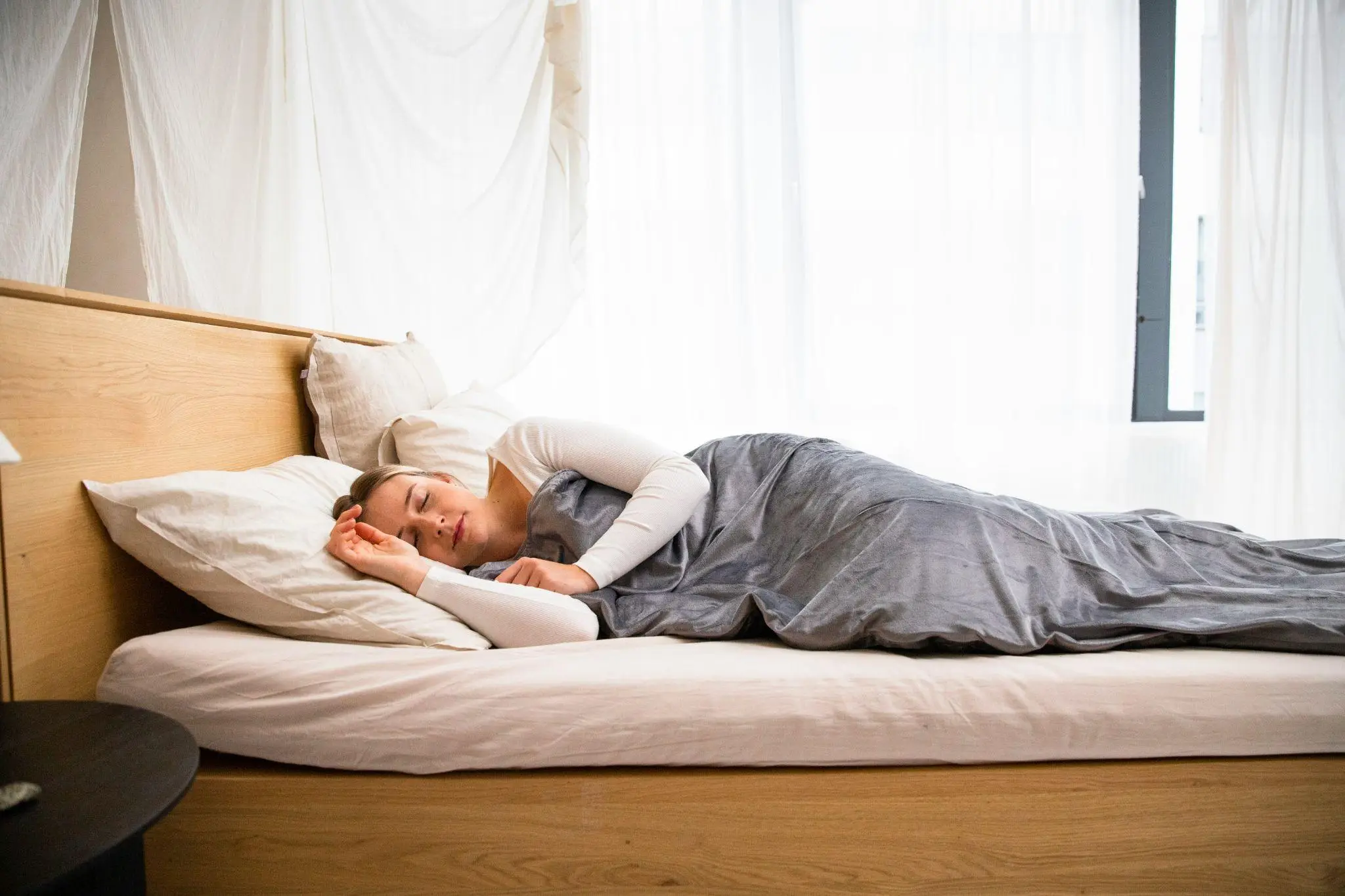 Person sleeping comfortably on a wooden bed with white and gray bedding in a bright room
