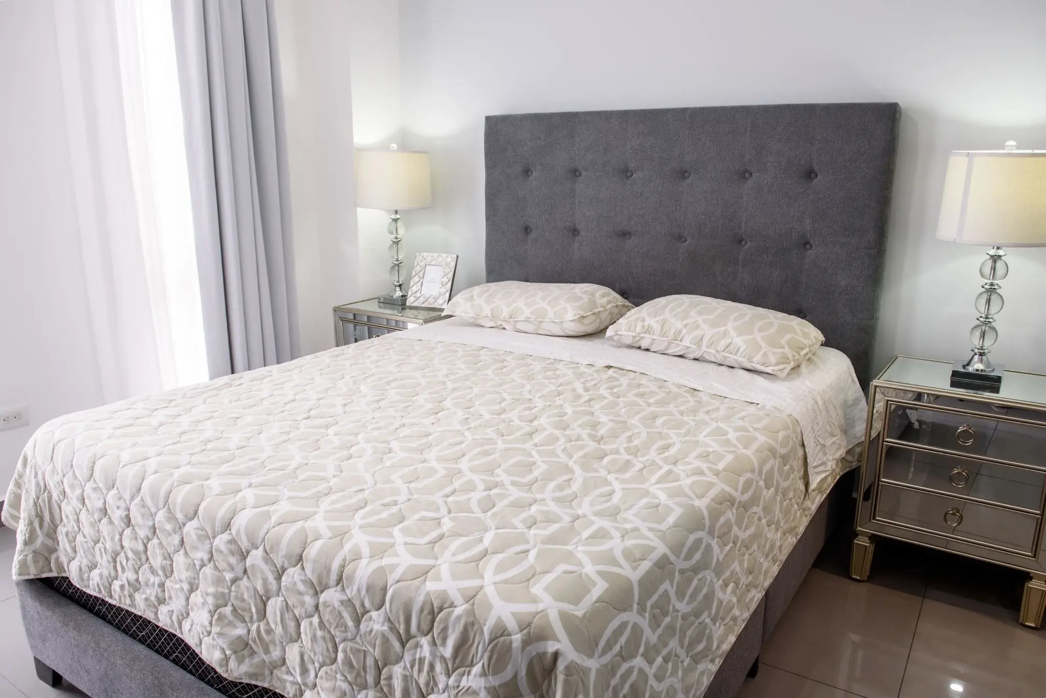Gray tufted headboard with patterned bedding in a bright bedroom featuring mirrored nightstands
