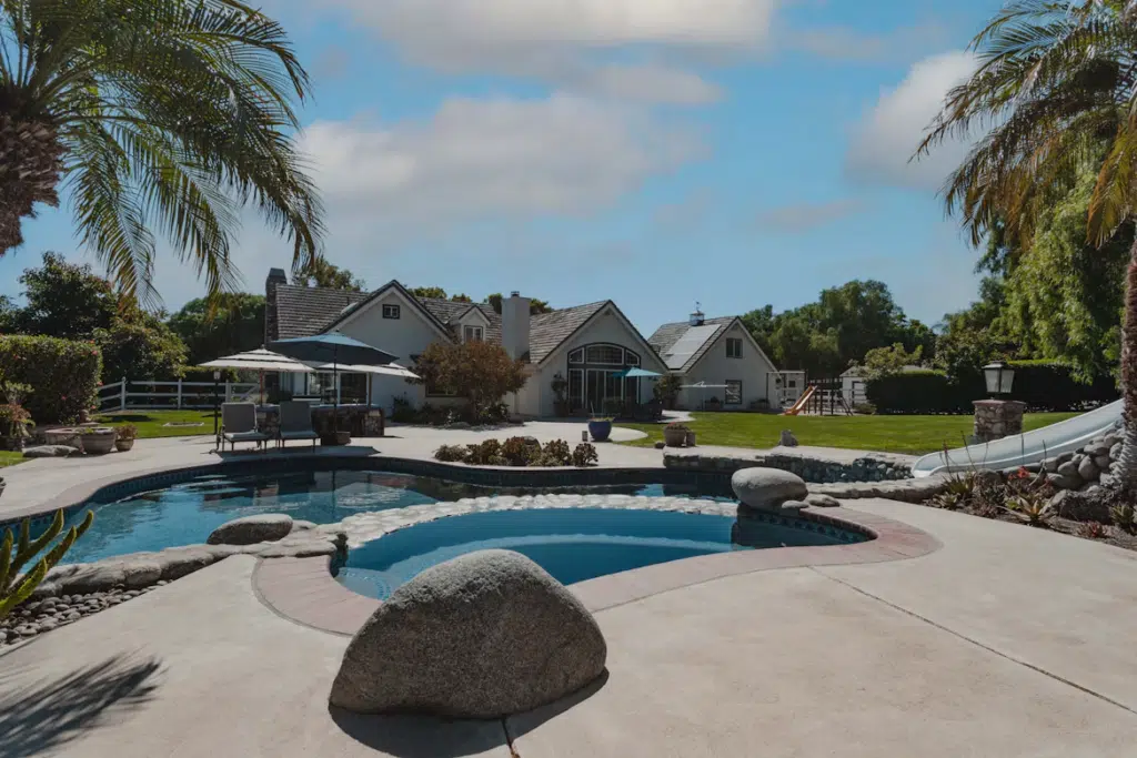 Backyard with pool, slide, and sun loungers beside large suburban house under blue sky