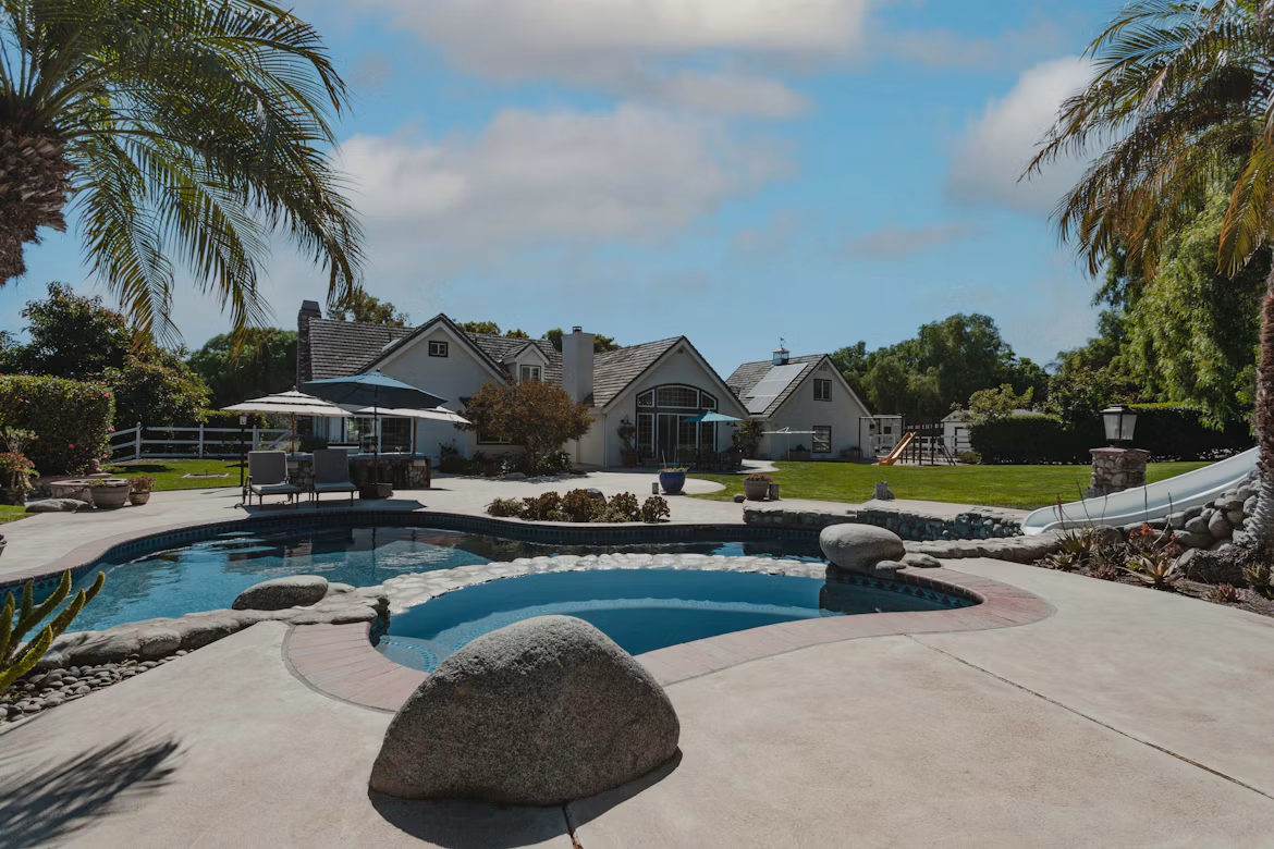 Backyard with pool, slide, and sun loungers beside large suburban house under blue sky