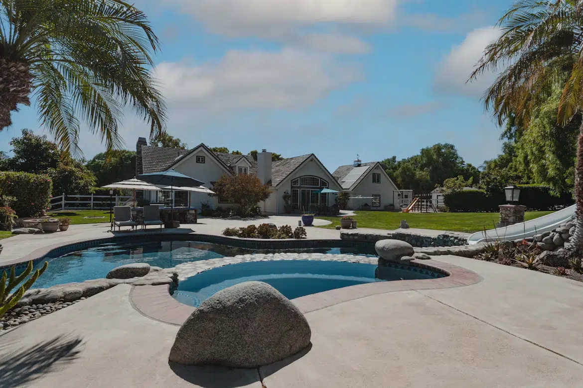 Backyard with pool, slide, and sun loungers beside large suburban house under blue sky