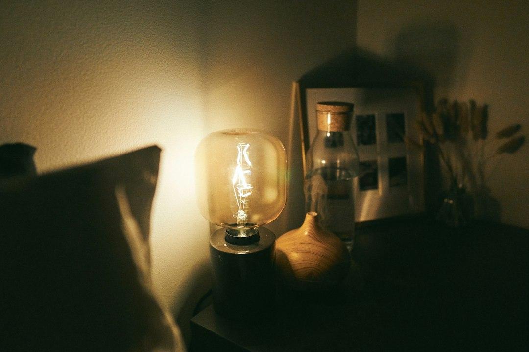 Antique-style bulb glowing on wooden nightstand with glass bottle and framed photo in dim light